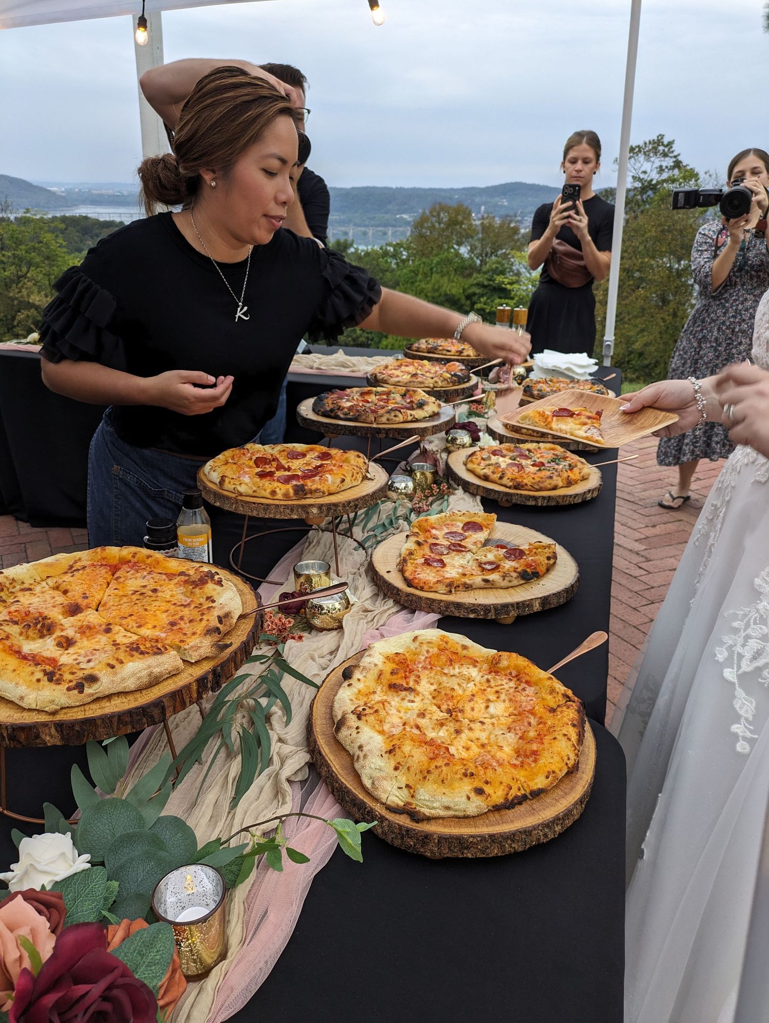 People gather around a table filled with freshly made pizzas on wooden boards at an outdoor event with mountain views.