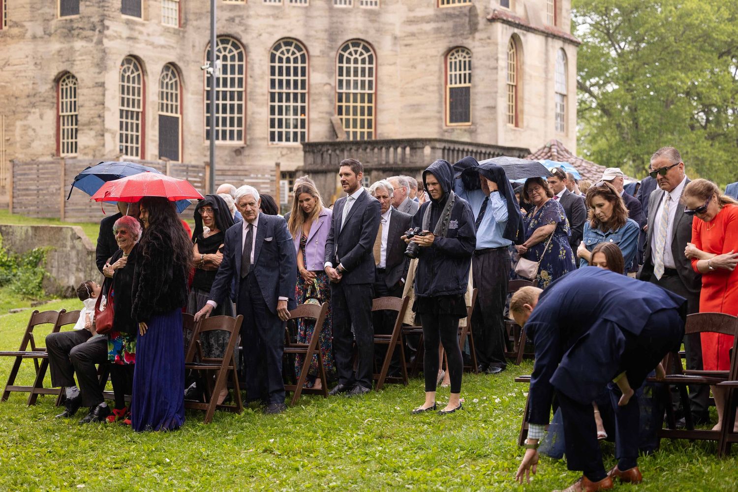 A group of formally dressed people gather under umbrellas on a rainy day outside a historic stone building.