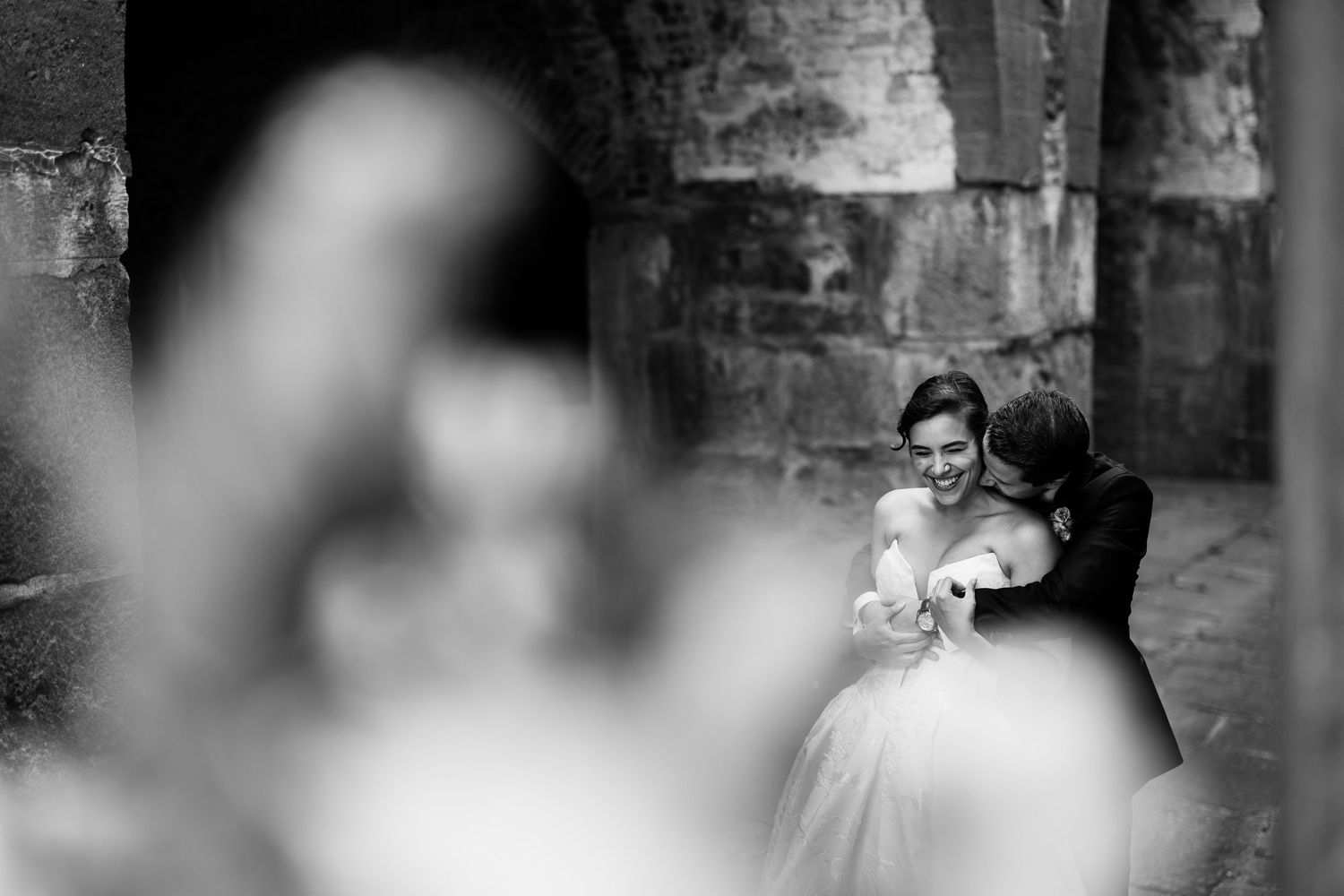 Romantic black and white portrait of newlyweds embracing near stone archway.