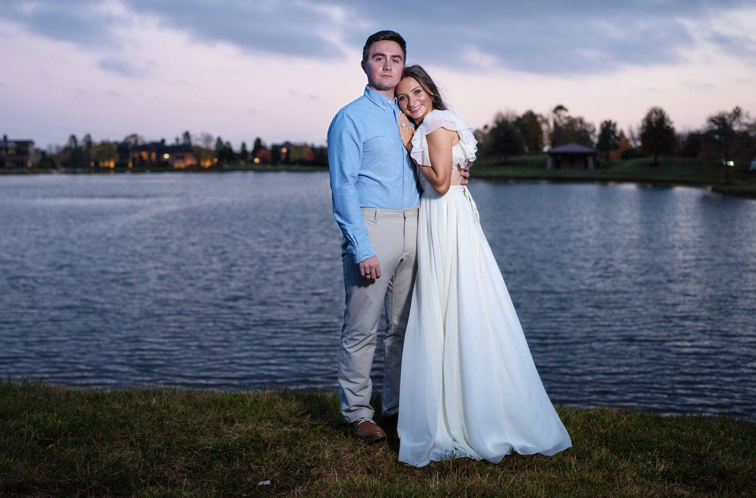A couple in light blue and white attire poses for romantic sunset photos by a serene lakeside.
