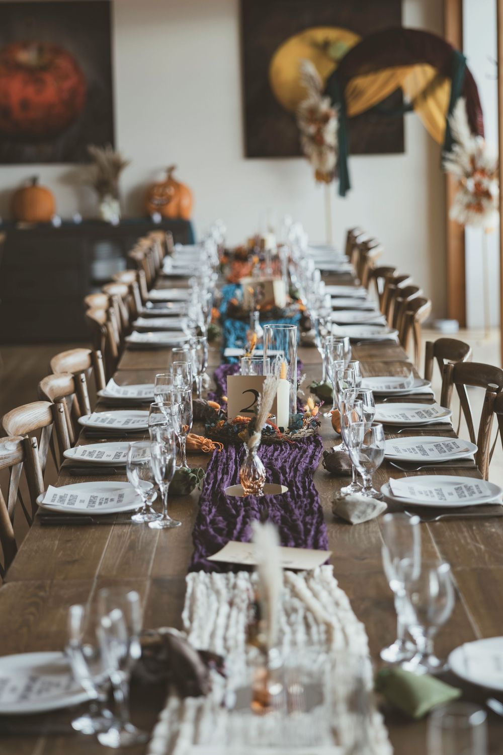 Long rustic wooden table set for an elegant dinner party with purple runner and white place settings.