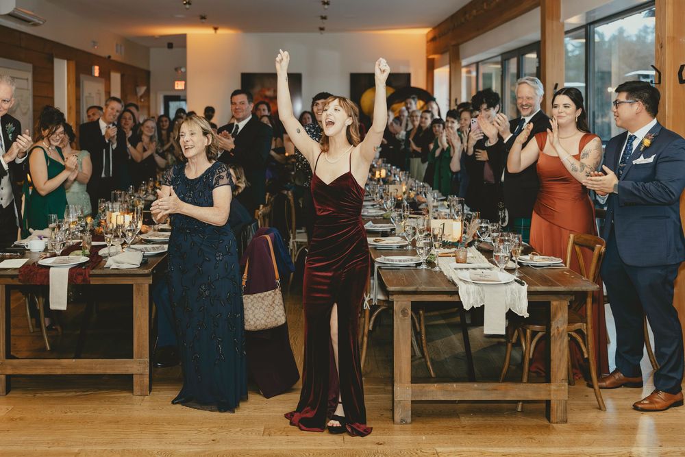 Wedding guests stand and applaud during a reception celebration at long wooden dining tables.