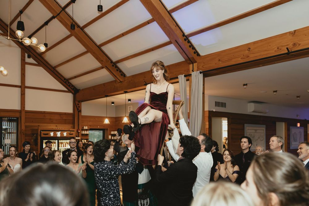 Wedding guests dancing and celebrating together in a rustic venue with exposed wooden beams and warm lighting.