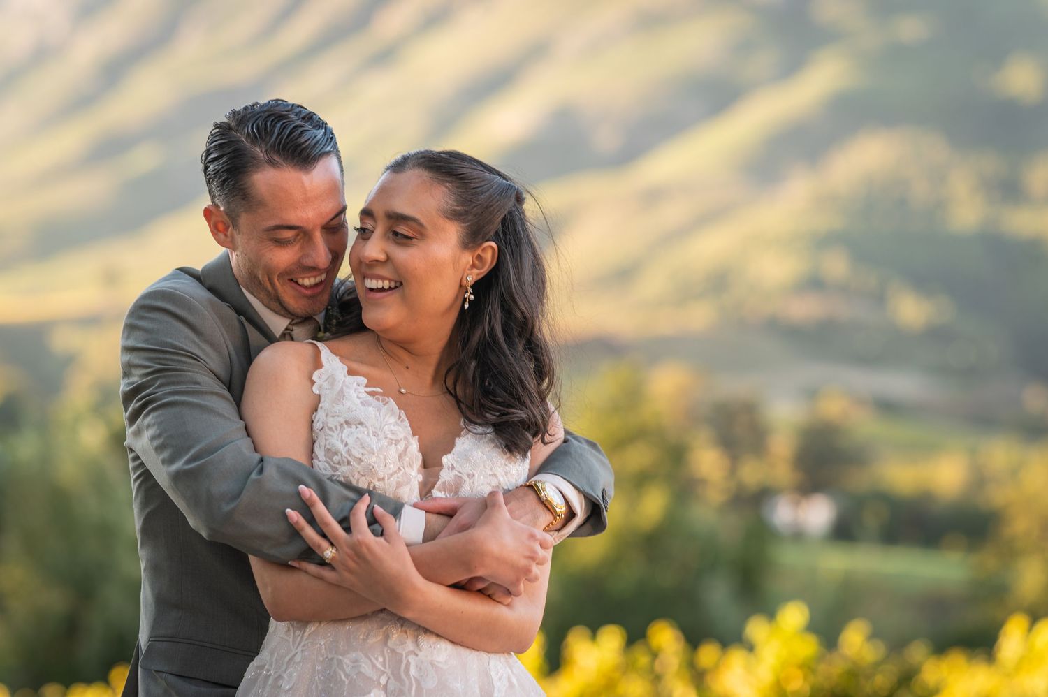 Happy couple embracing outdoors in wedding attire with scenic green mountains in golden sunlight background.