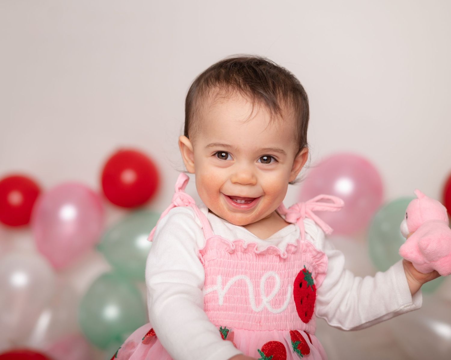 Smiling baby in white and pink outfit sits among red, pink and mint colored party balloons.