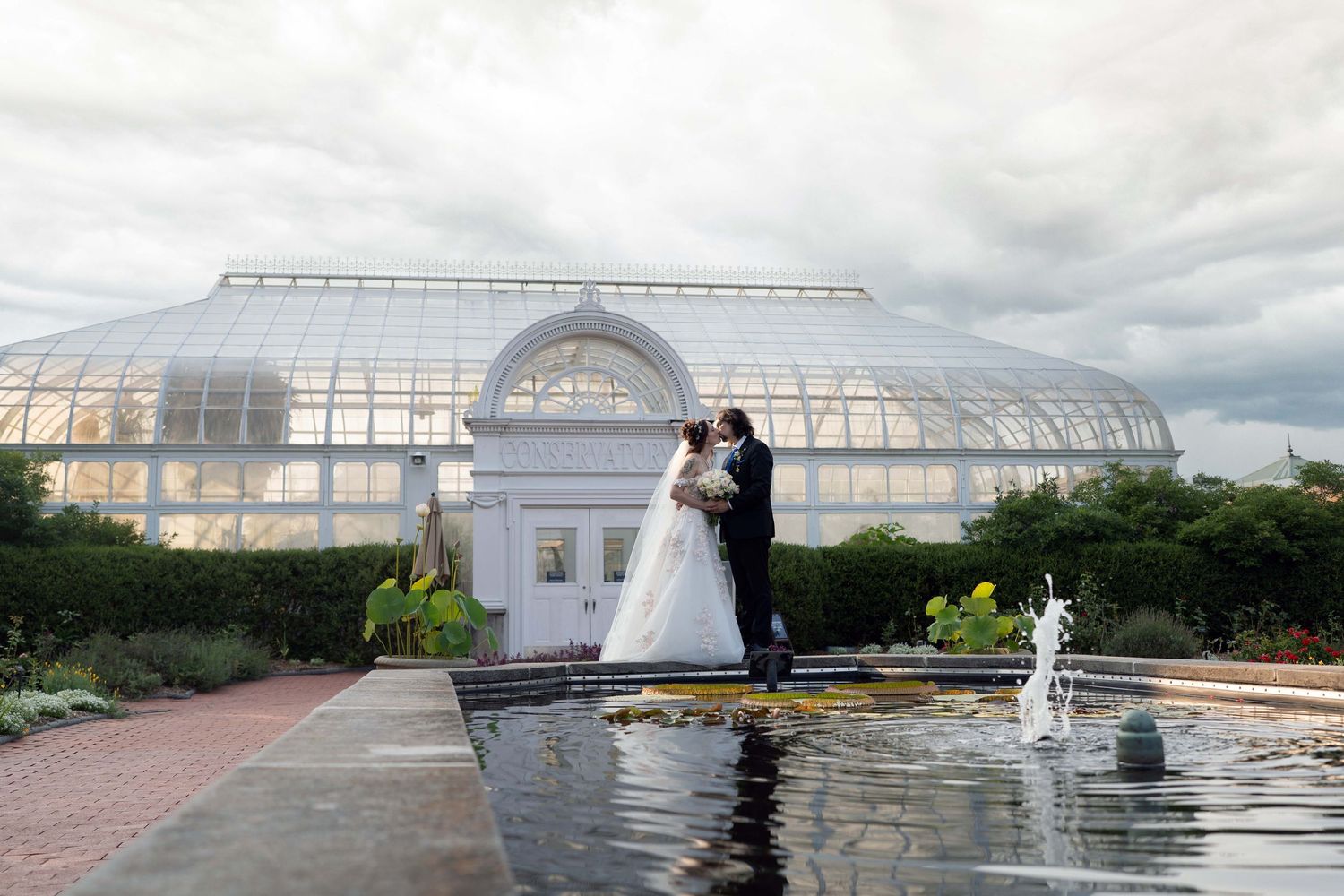 Bride and groom kissing near a fountain outside a grand glass greenhouse wedding venue at Toledo Zoo Wedding Venue