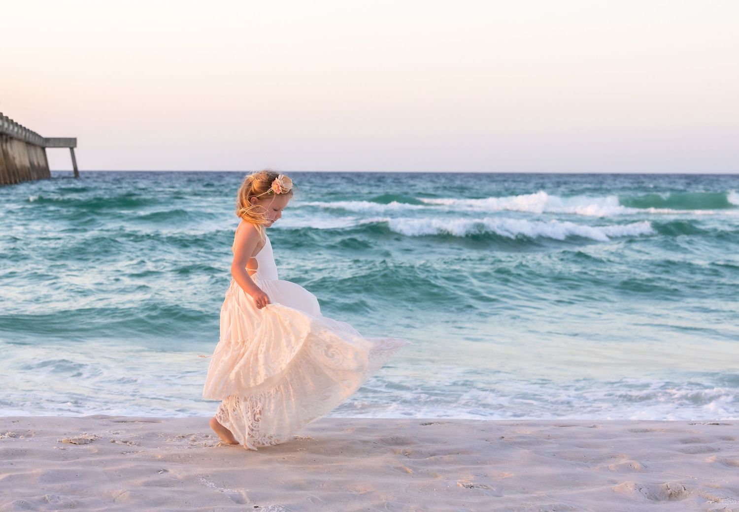 Young girl in white dress and hat twirling on a sandy beach with turquoise ocean waves at sunset.