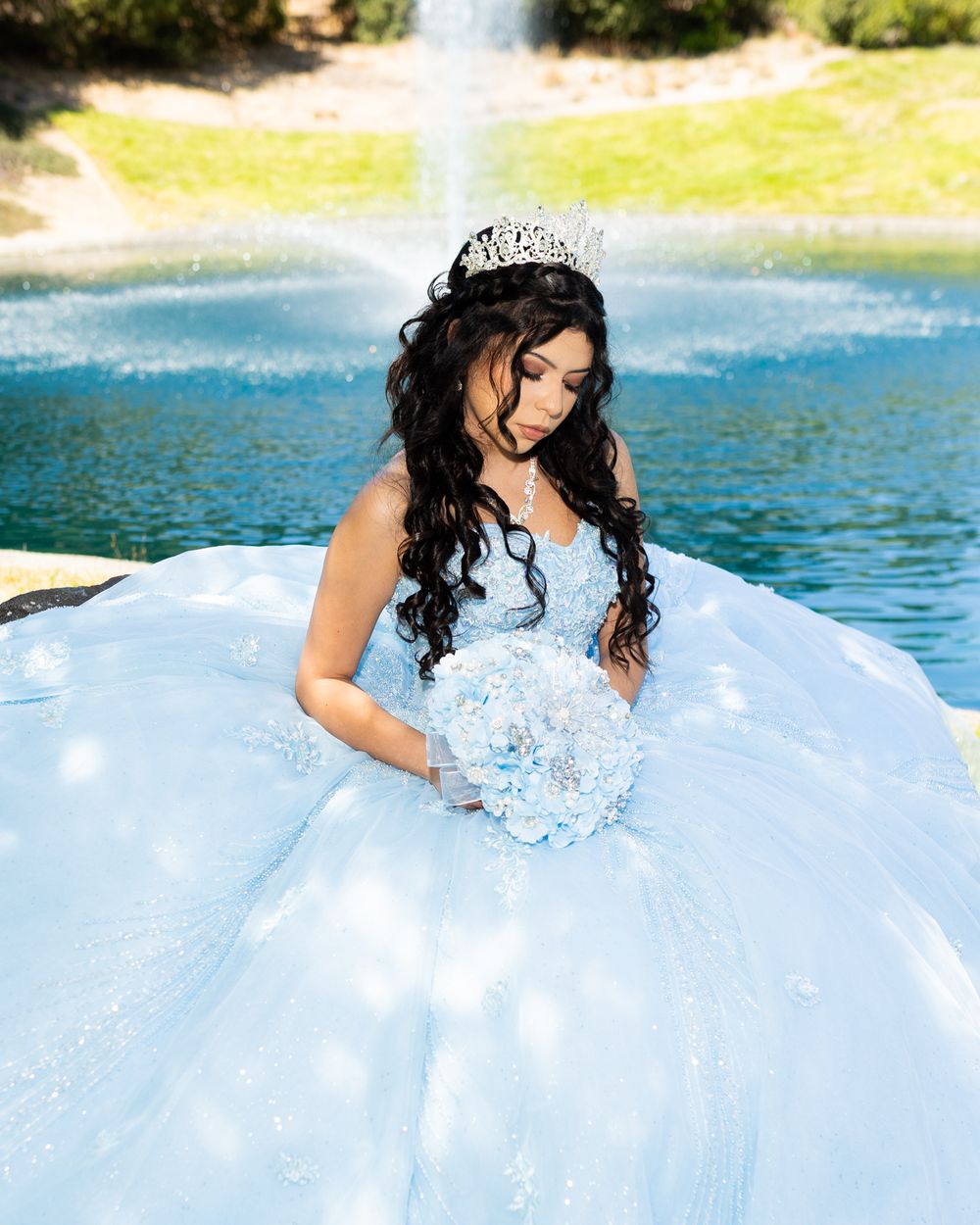Young woman in light blue quinceañera ball gown with tiara holds floral bouquet near fountain.