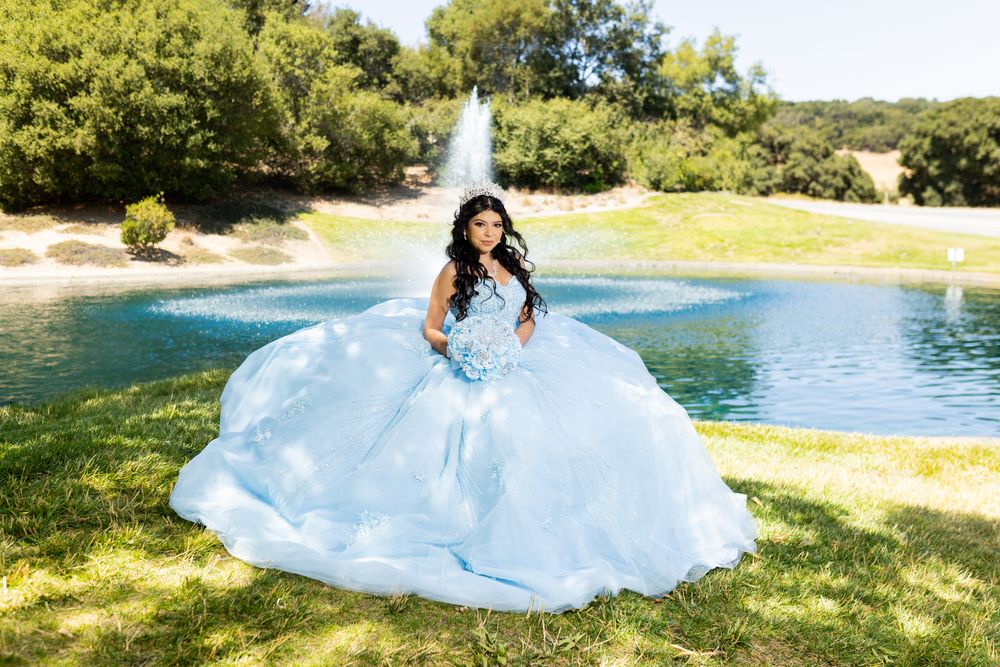 Young woman in a voluminous light blue quinceañera ball gown poses by a fountain in a lush green park.