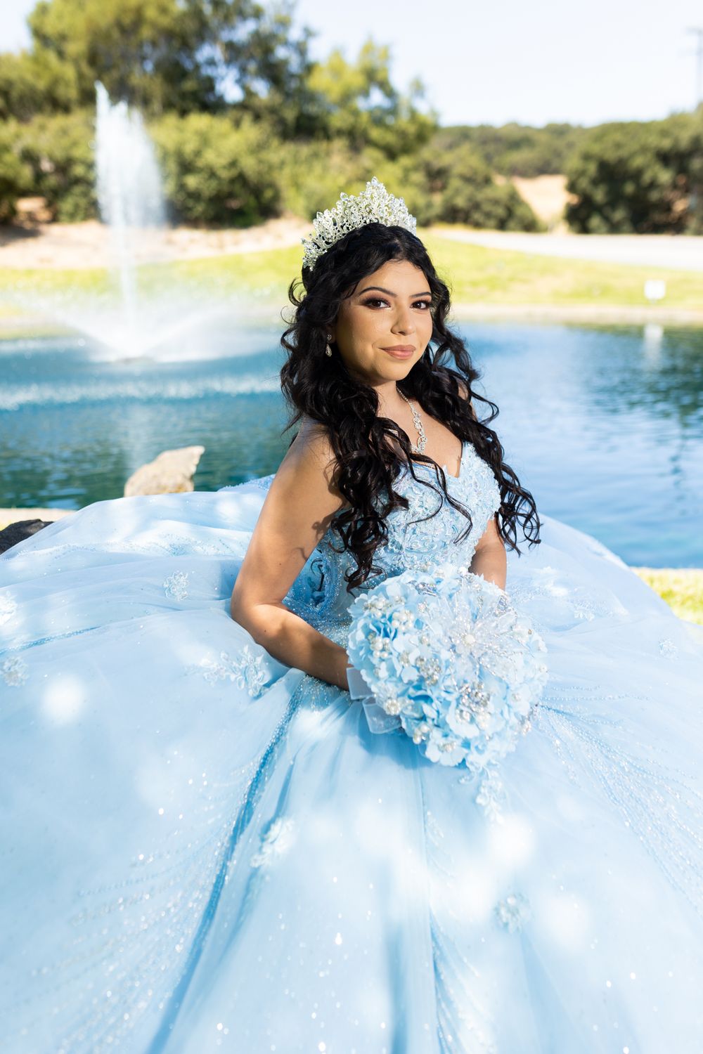 Young woman in light blue quinceañera ball gown with tiara and bouquet poses by fountain.