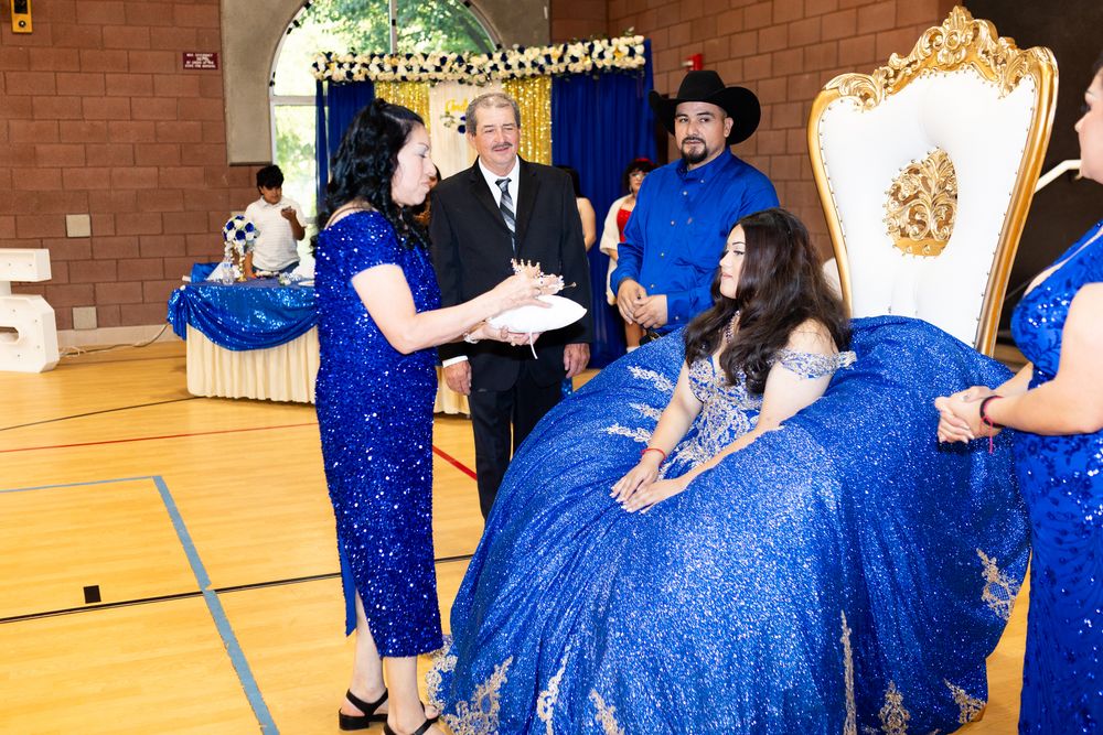 A quinceañera celebration in a gym with guests in royal blue sequin gowns and a cowboy-hatted man near a golden throne.