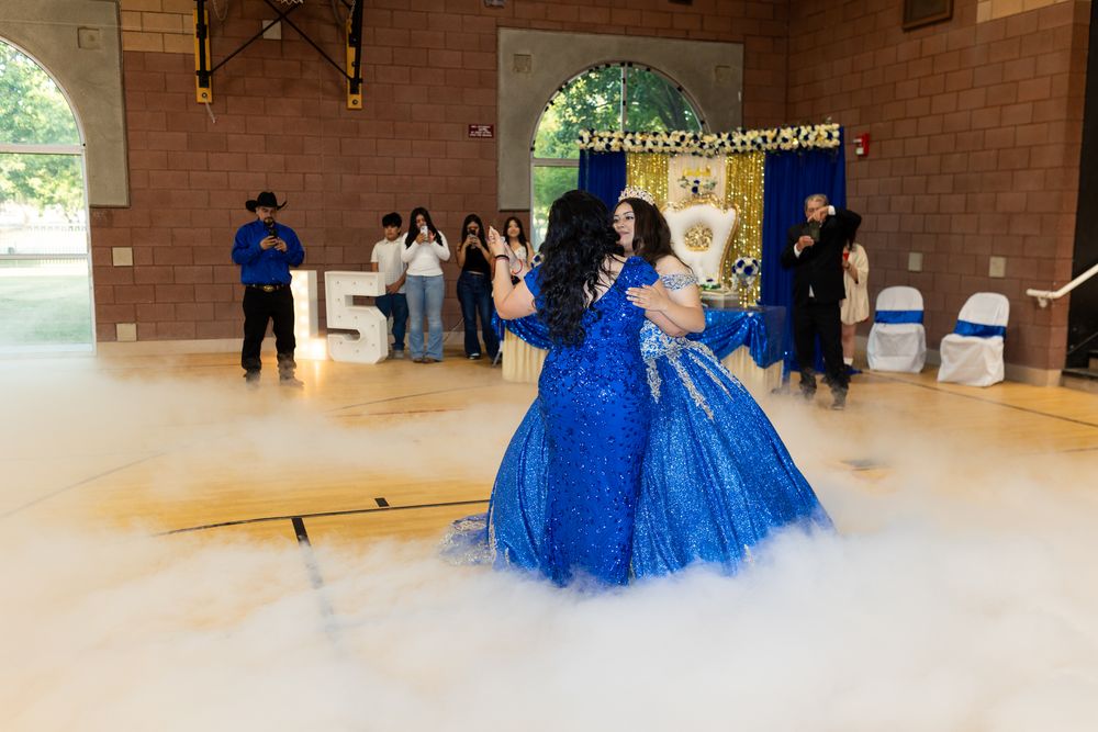 Two women in blue quinceañera gowns dance on a foggy floor at a 15th birthday celebration.