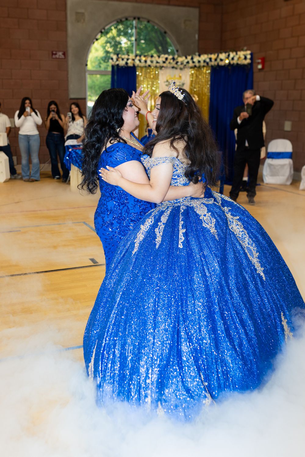 Two women in matching royal blue sequin ball gowns share a dance at a quinceañera celebration.