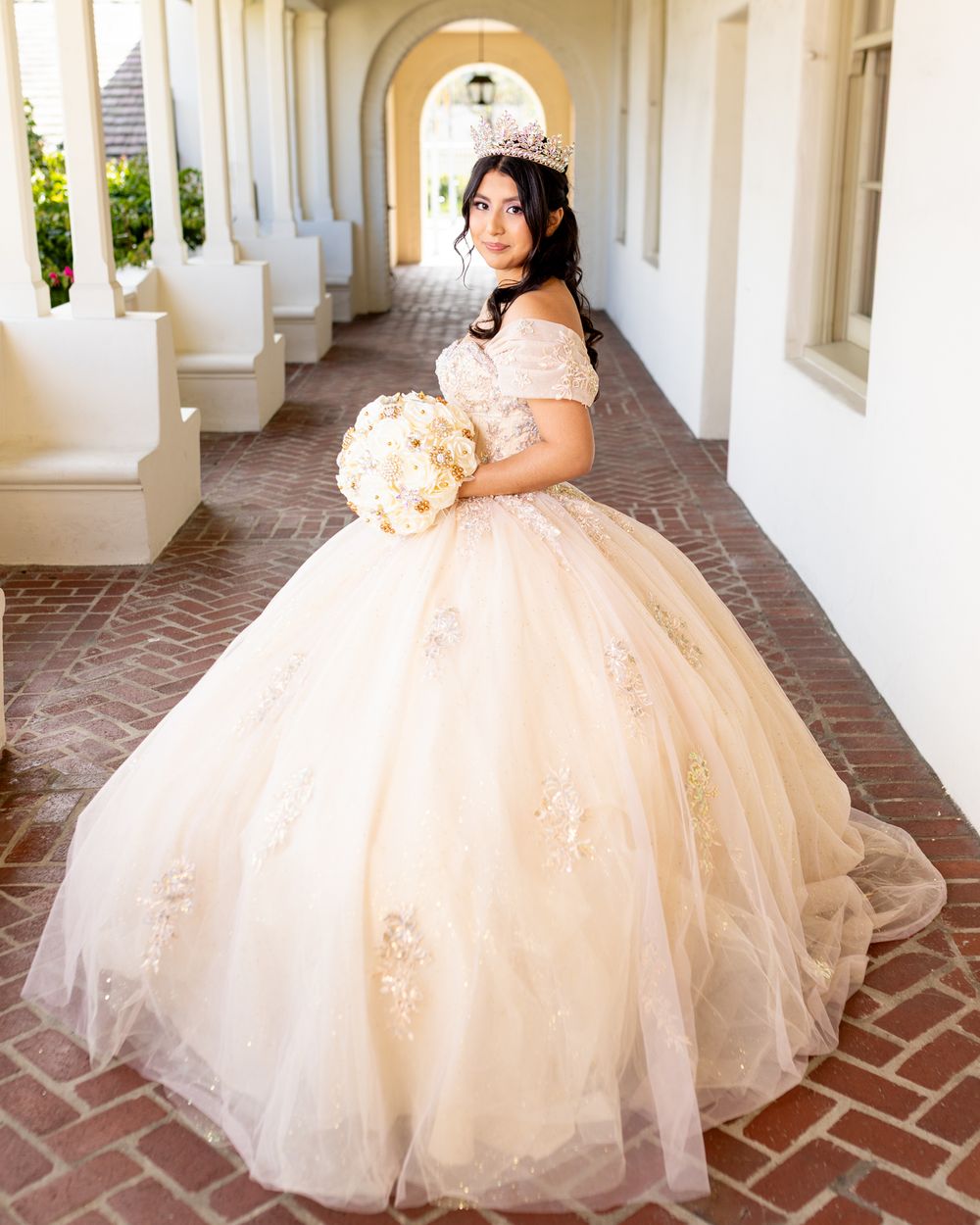 Young woman in champagne ball gown with floral appliques and tiara holds white bouquet in elegant archway.