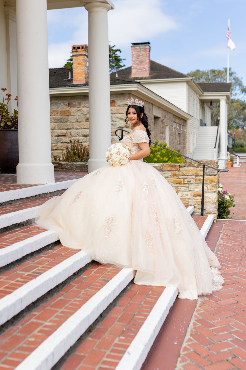 Young woman in a blush ballgown and tiara holds a bouquet on brick steps outside a stone venue.