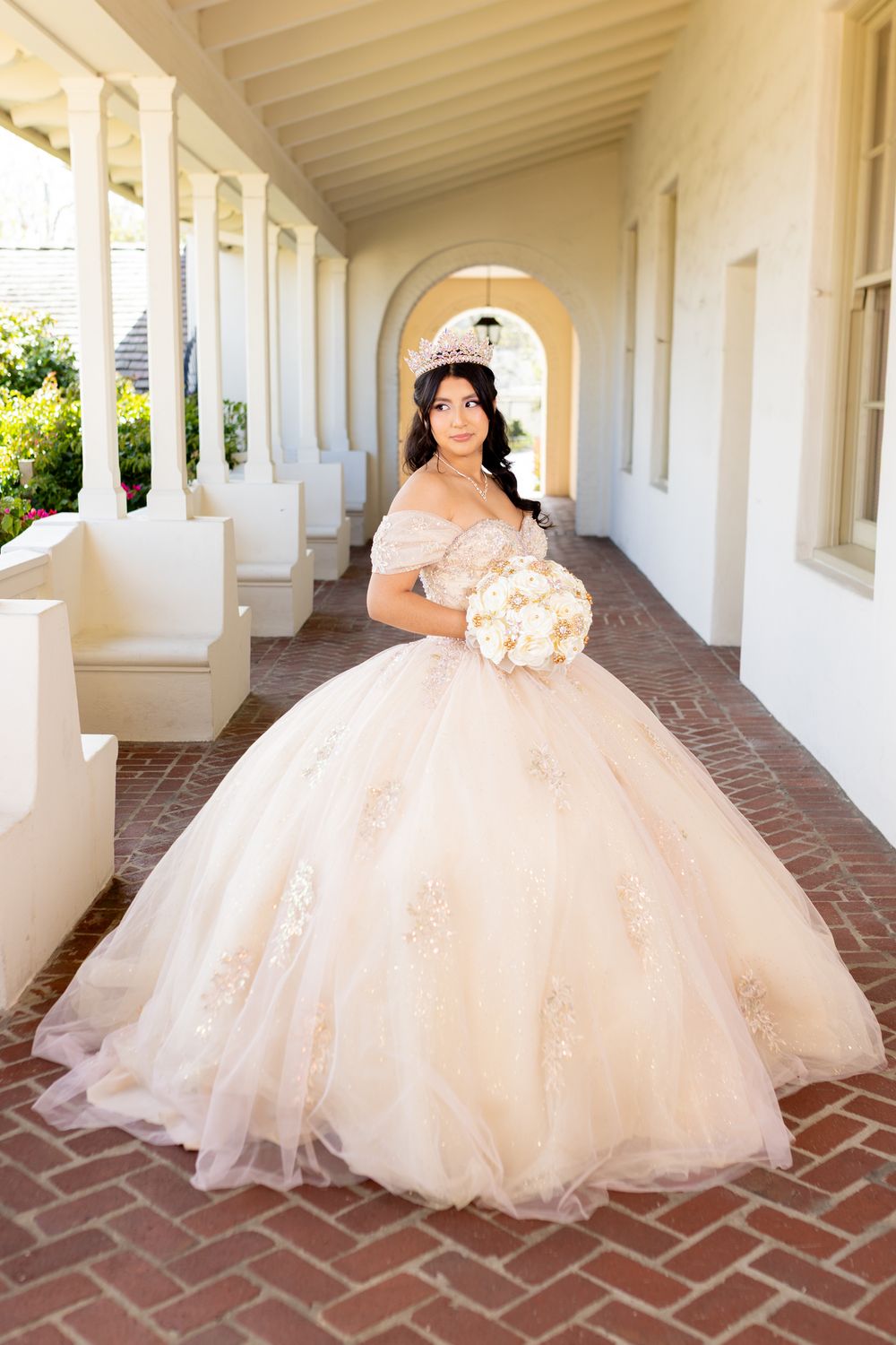 Young woman in blush pink quinceañera ball gown with tiara holds bouquet in elegant arched corridor.