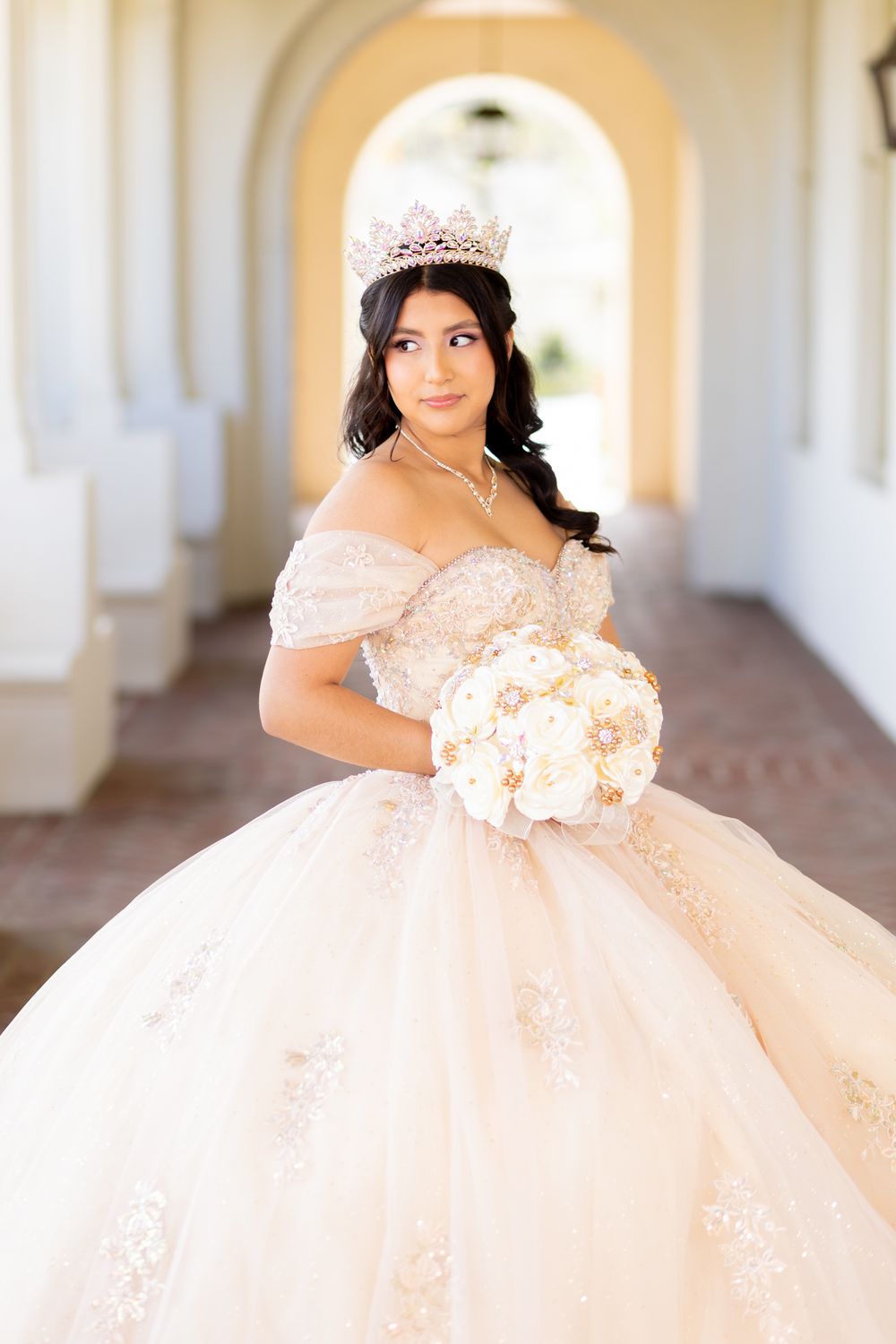 Young woman in elegant blush ball gown with tiara holds white bouquet in sunlit archway corridor.