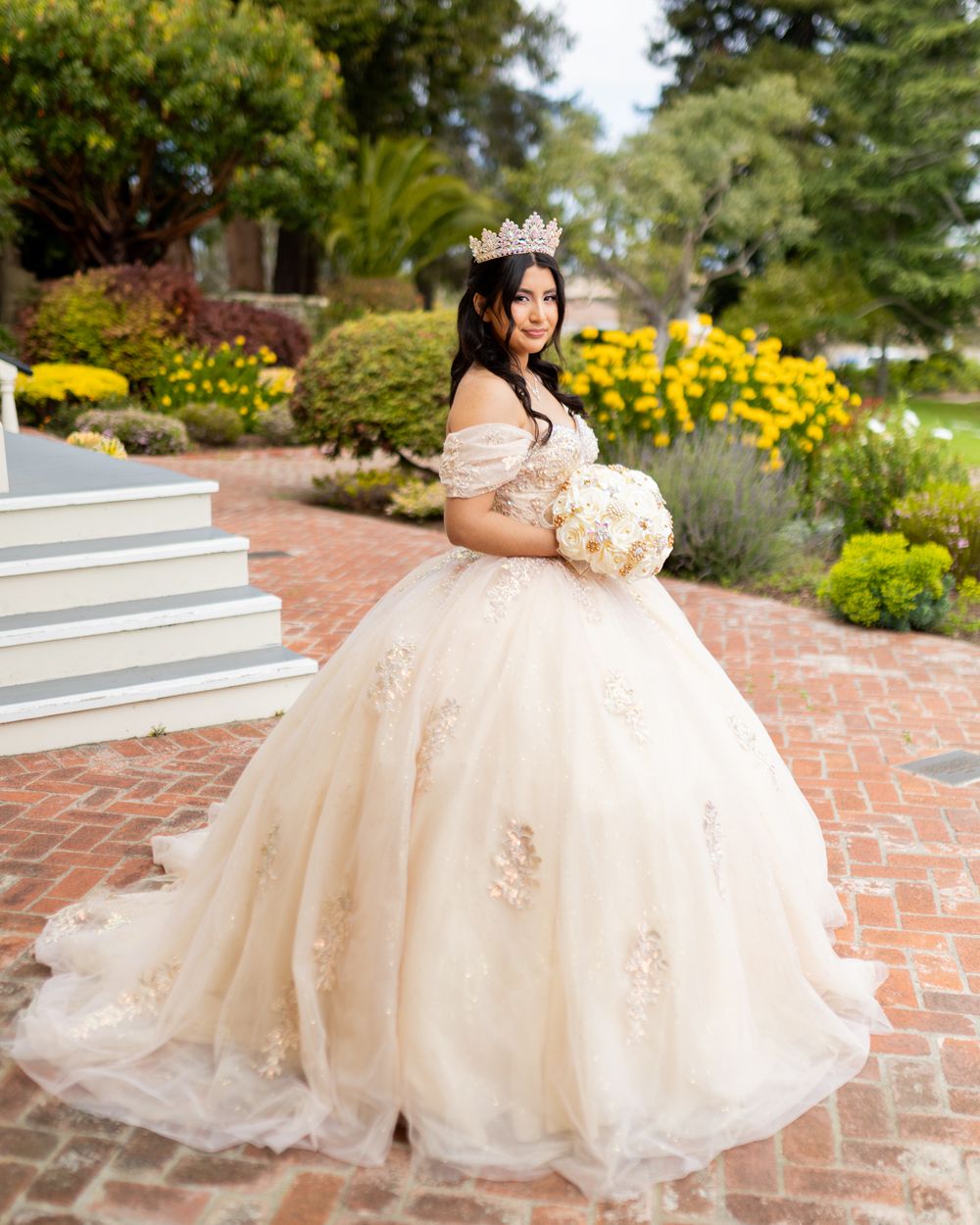 Young woman in elegant champagne ball gown with floral appliques and tiara holds bouquet in garden.