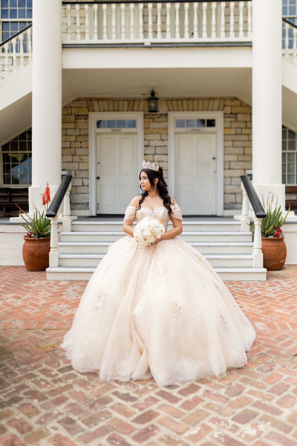 Young woman in blush ball gown and tiara holds white bouquet on brick steps of elegant stone mansion.