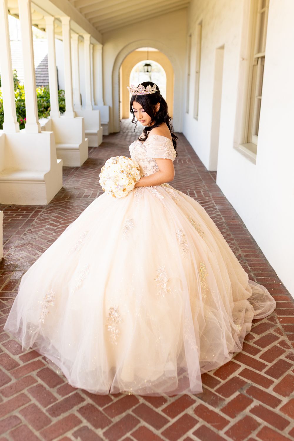 Young woman in elegant champagne ball gown with tiara and bouquet poses in archway for quinceañera.