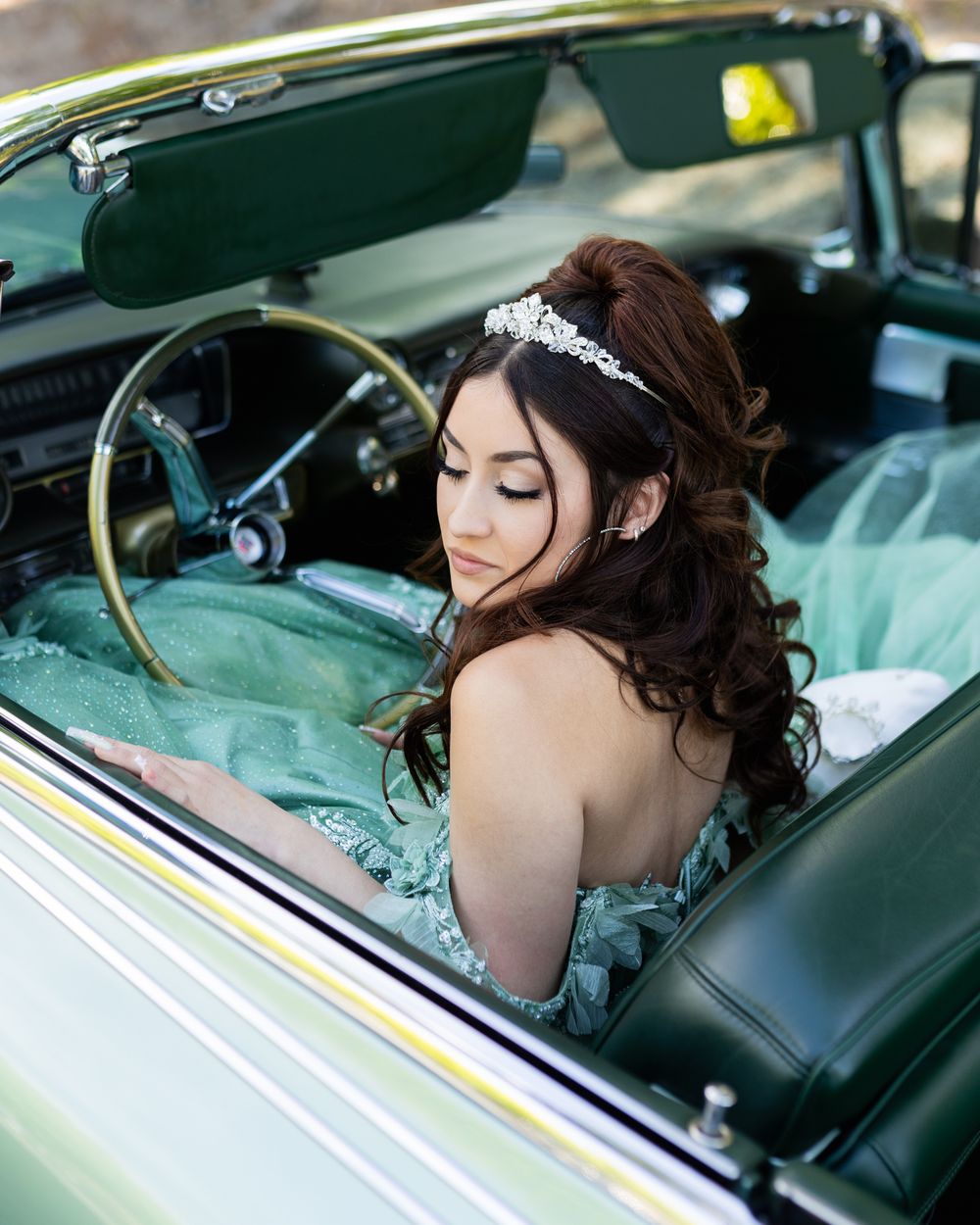 Young woman in mint green ball gown and tiara sitting in a vintage green convertible car for quinceañera photos.