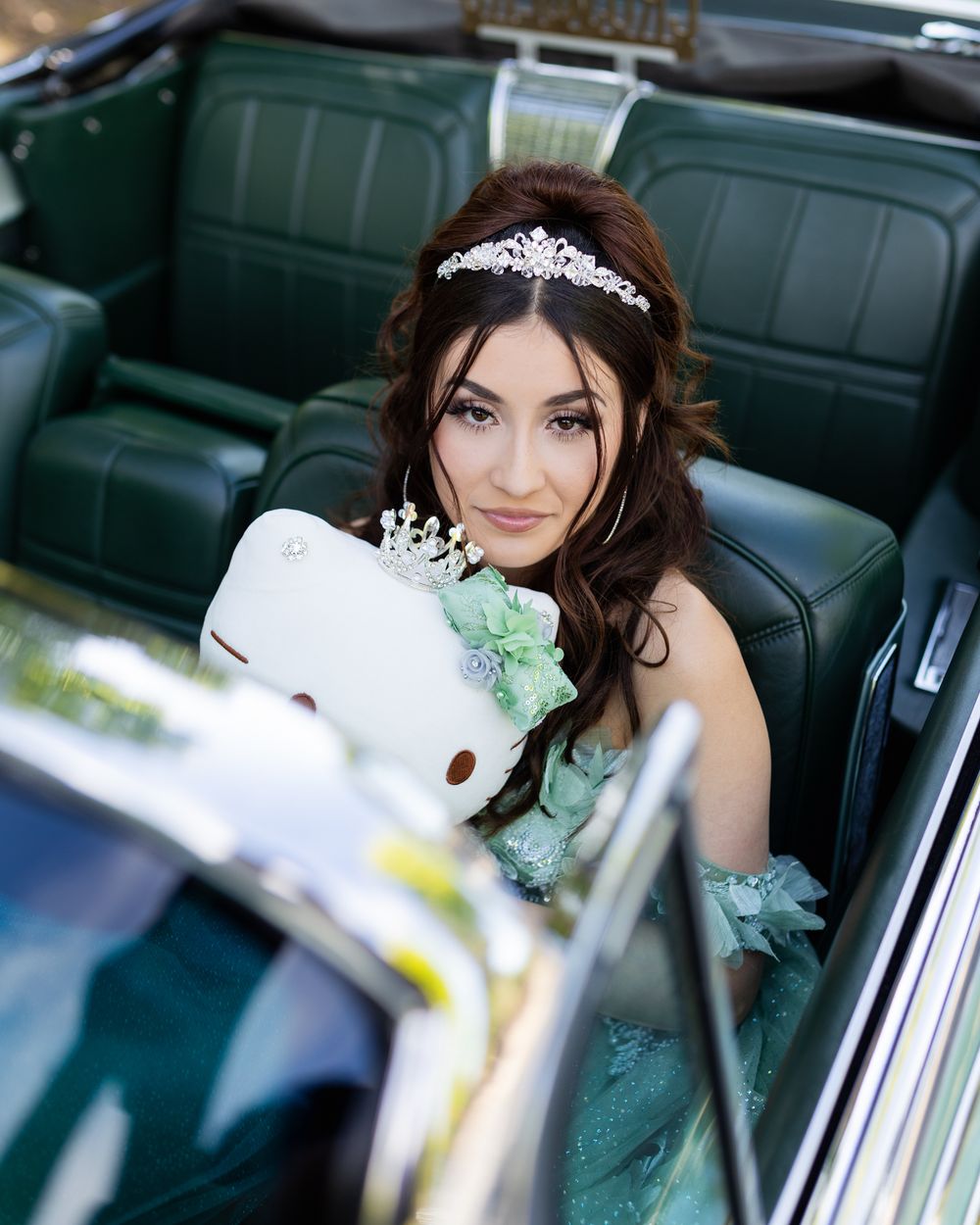 Young woman in a green gown and tiara holds a Hello Kitty plush inside a vintage green convertible car.