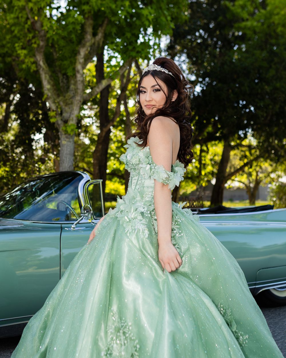 Young woman in a mint green ball gown with floral details and tiara poses by a vintage convertible car.