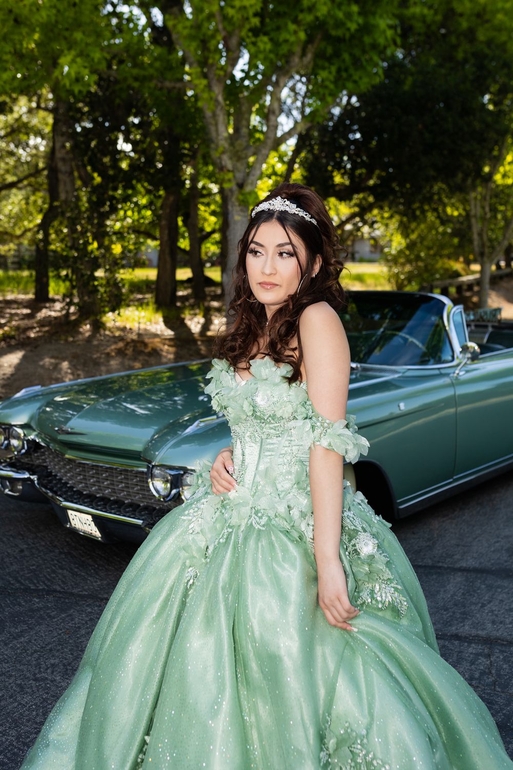 Young woman in mint green quinceañera ball gown with tiara poses beside a vintage teal convertible car outdoors.