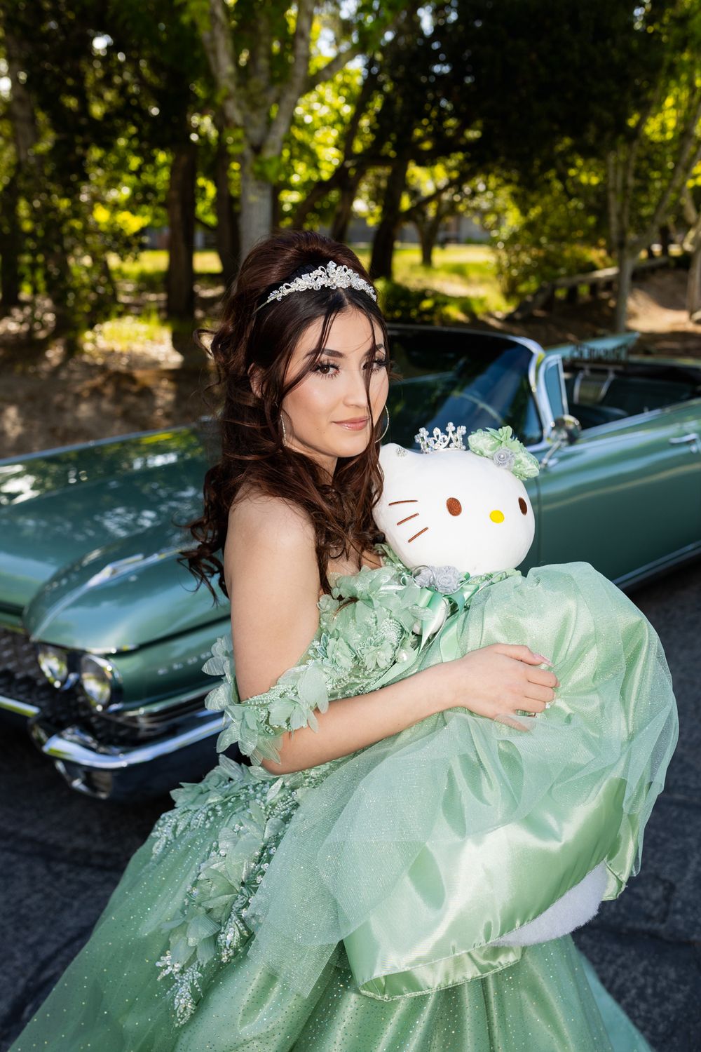 Young woman in mint green quinceañera gown holding Hello Kitty plush beside a vintage teal convertible.