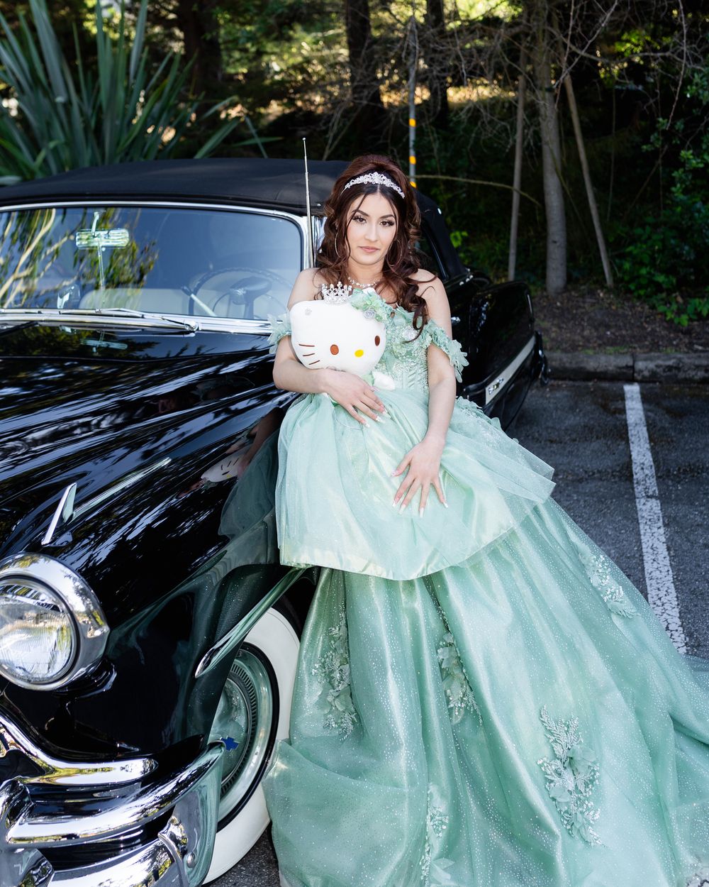 Young woman in mint green quinceañera gown holding Hello Kitty plush beside a classic black vintage car outdoors.