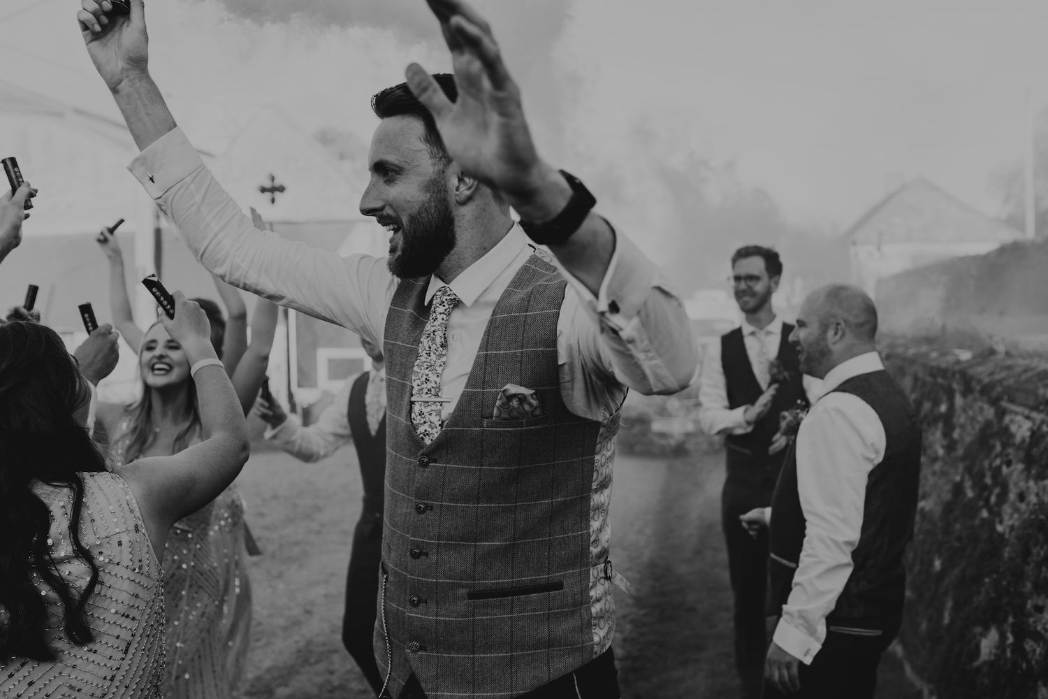 Black and white photo of a groom celebrating outdoors with guests at a wedding reception with smoke effects.