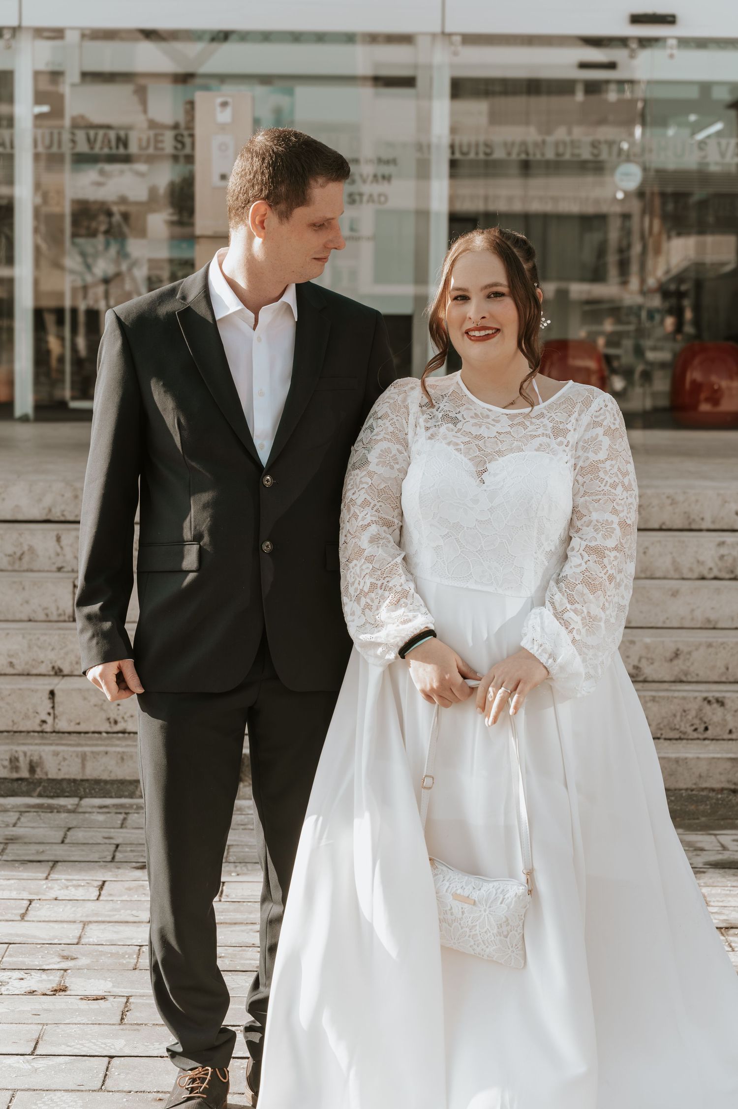 A bride in a white lace gown and groom in a black suit hold hands on steps outside a building.