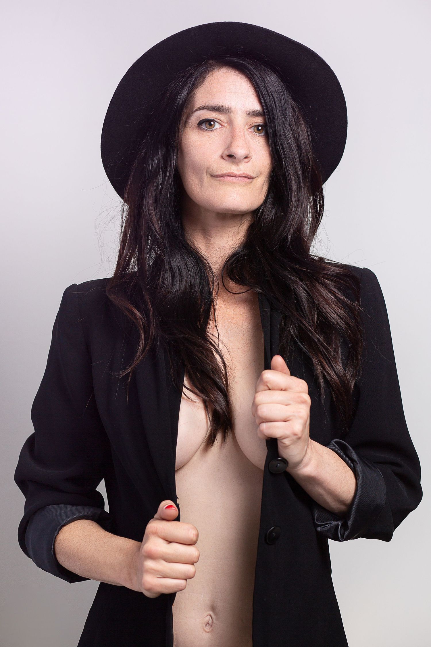 Woman in black blazer and wide-brim hat posing confidently against a light gray studio background.