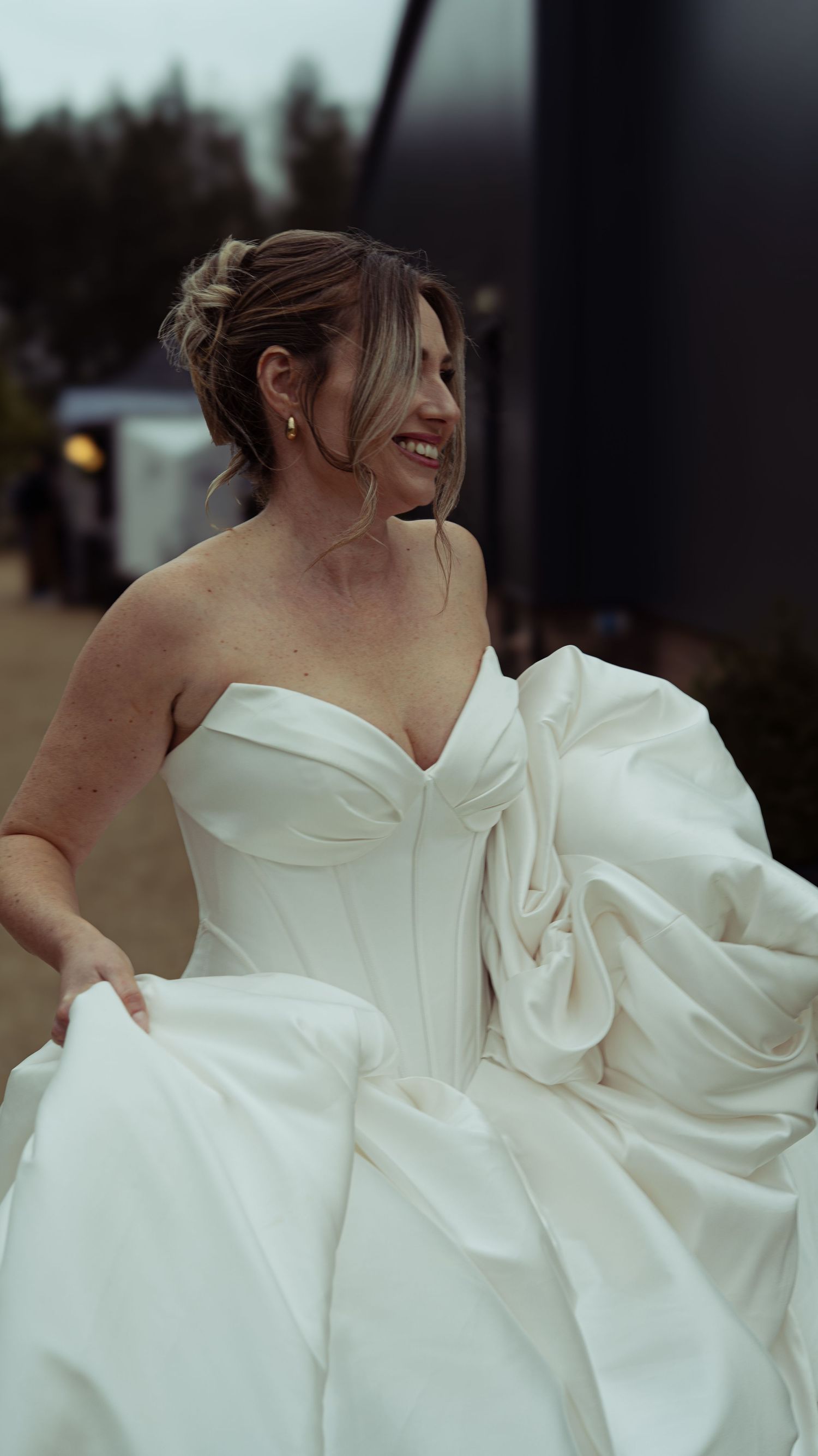 Smiling bride in a strapless white ball gown with dramatic bow detail, holding her dress outdoors.