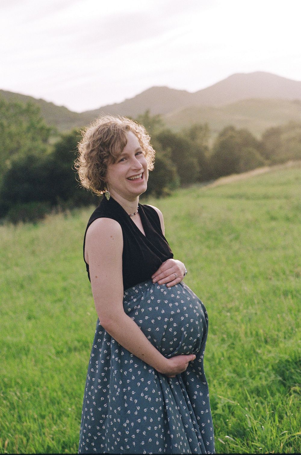 Film maternity photography in California with green rolling hills behind a pregnant woman dancing in the grass.