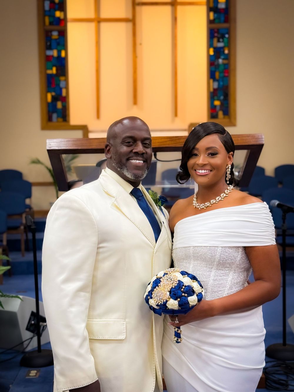 Smiling bride and groom in white wedding attire pose together in a church, bride holding blue and gold bouquet.
