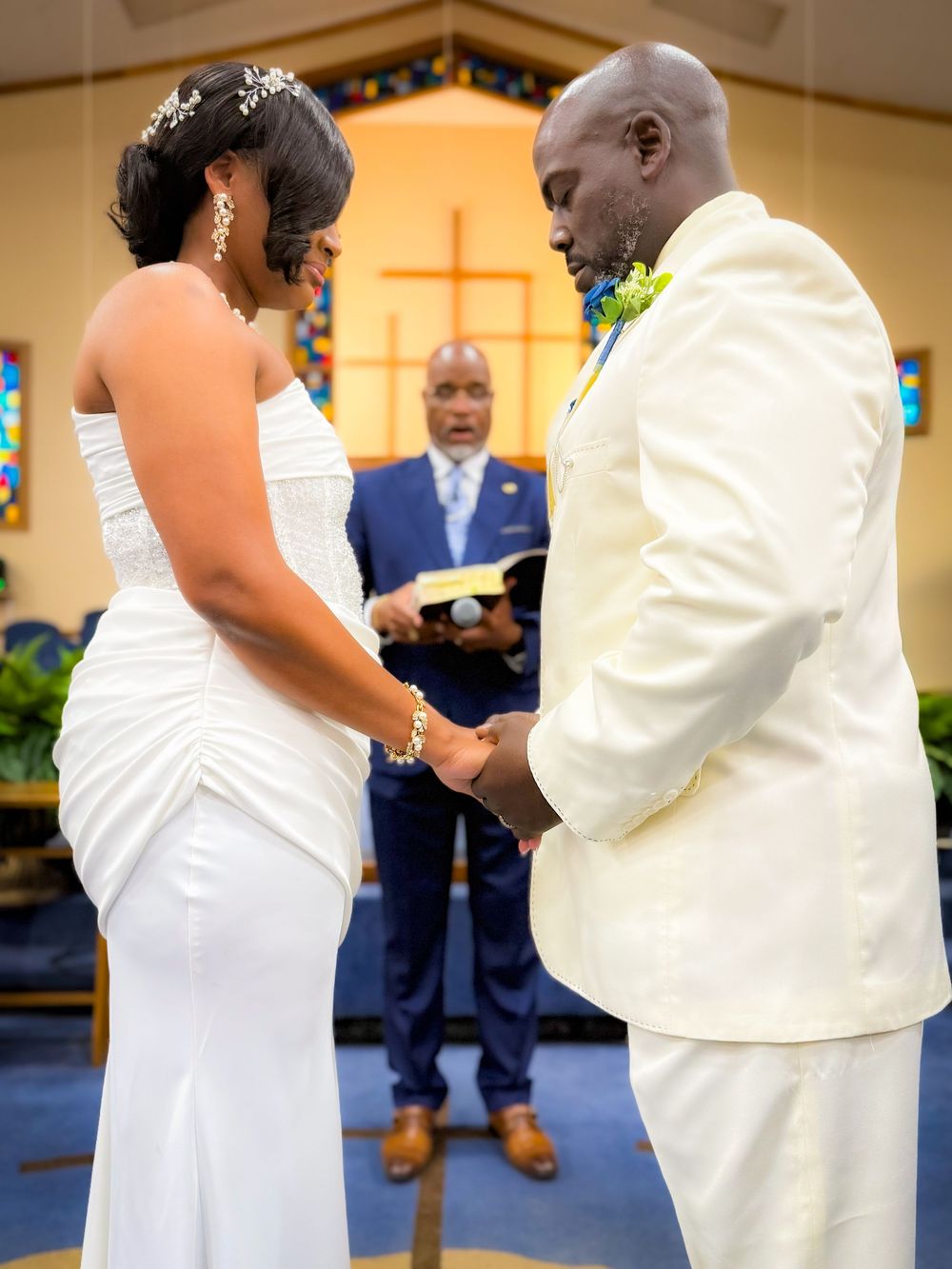 Bride and groom hold hands at church altar during wedding ceremony, officiant reads vows in background.