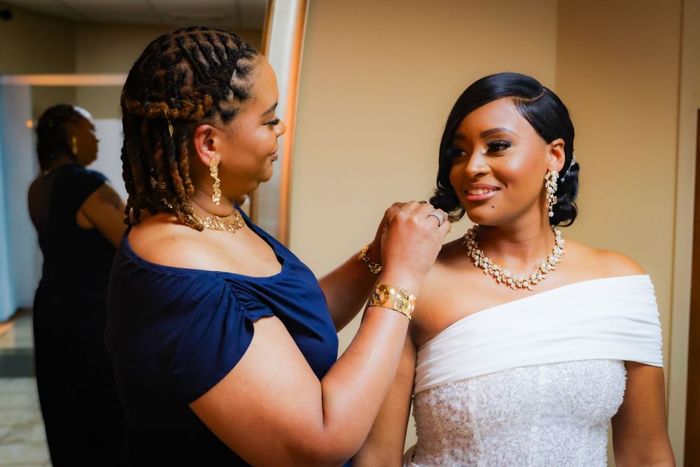 Woman in navy dress helps bride in white off-shoulder gown with gold necklace during wedding preparations.