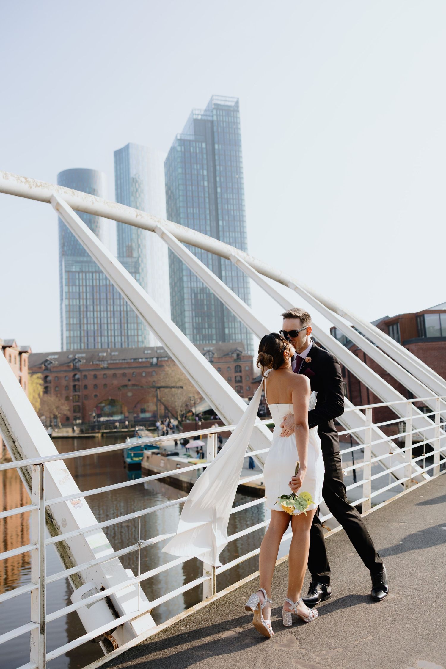Smiling couple in sunglasses posing on a rooftop terrace, groom in black suit, bride in white mini dress, urban bridge backdrop.