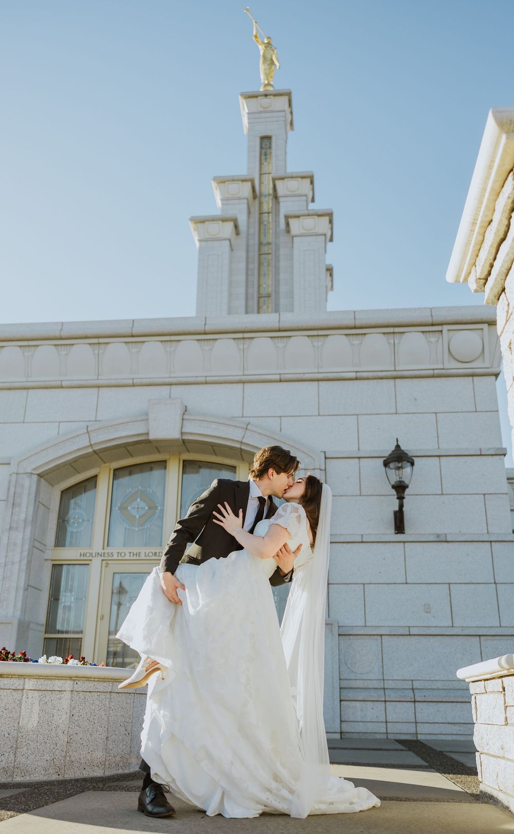 A groom dips his bride in front of a white bricked chapel