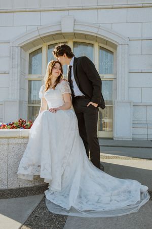 A groom kisses his bride on the top of her head. She is sitting on a concrete flower bed.