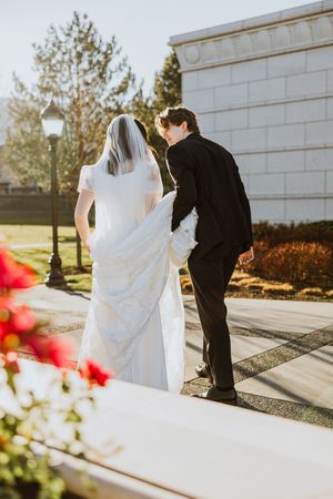 A bride and groom walk away into the sun. The groom is holding her dress train and there are pink flowers in the foreground