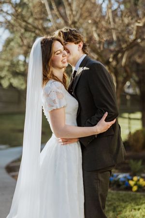 A man kisses his wife on her cheek while she laughs, they are in wedding attire