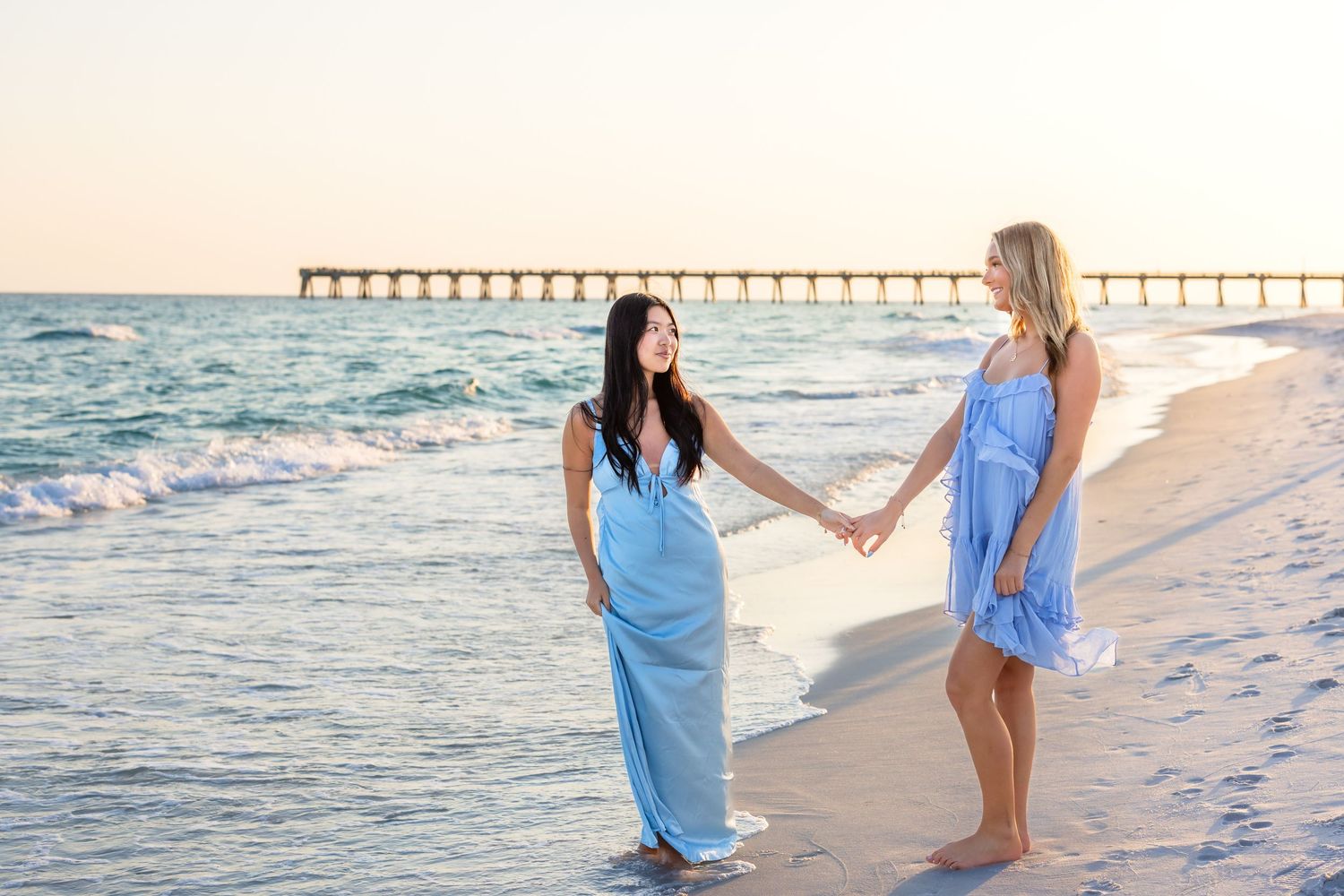 Two women in blue dresses embrace on a beach at sunset with a pier in the background.