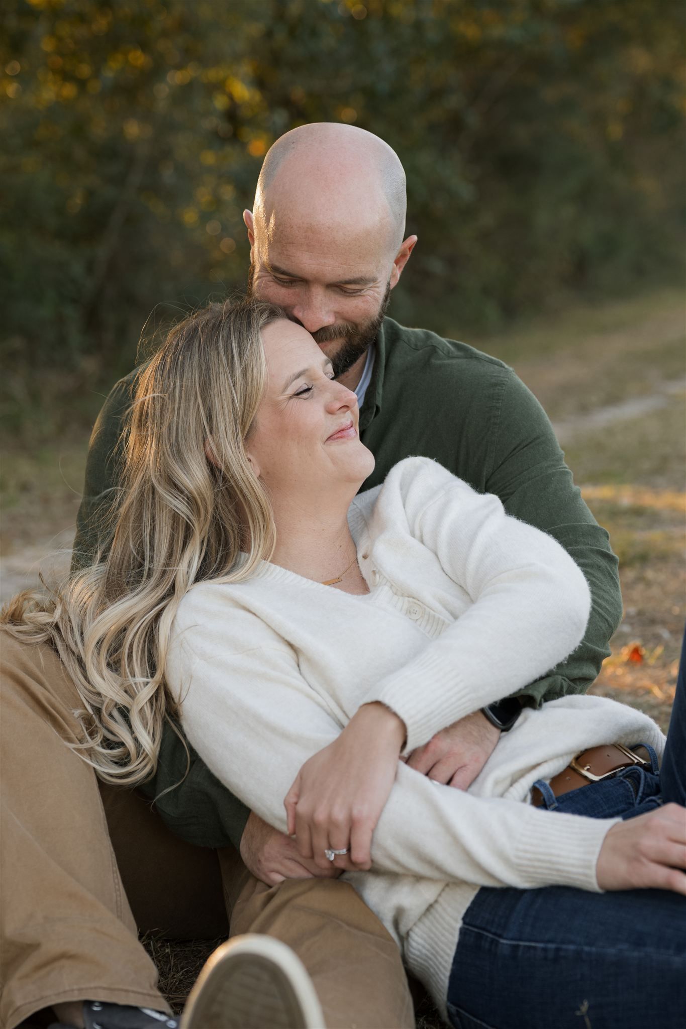 Couple sitting outdoors in autumn, woman in white sweater leaning back on man in green sweater, smiling warmly.