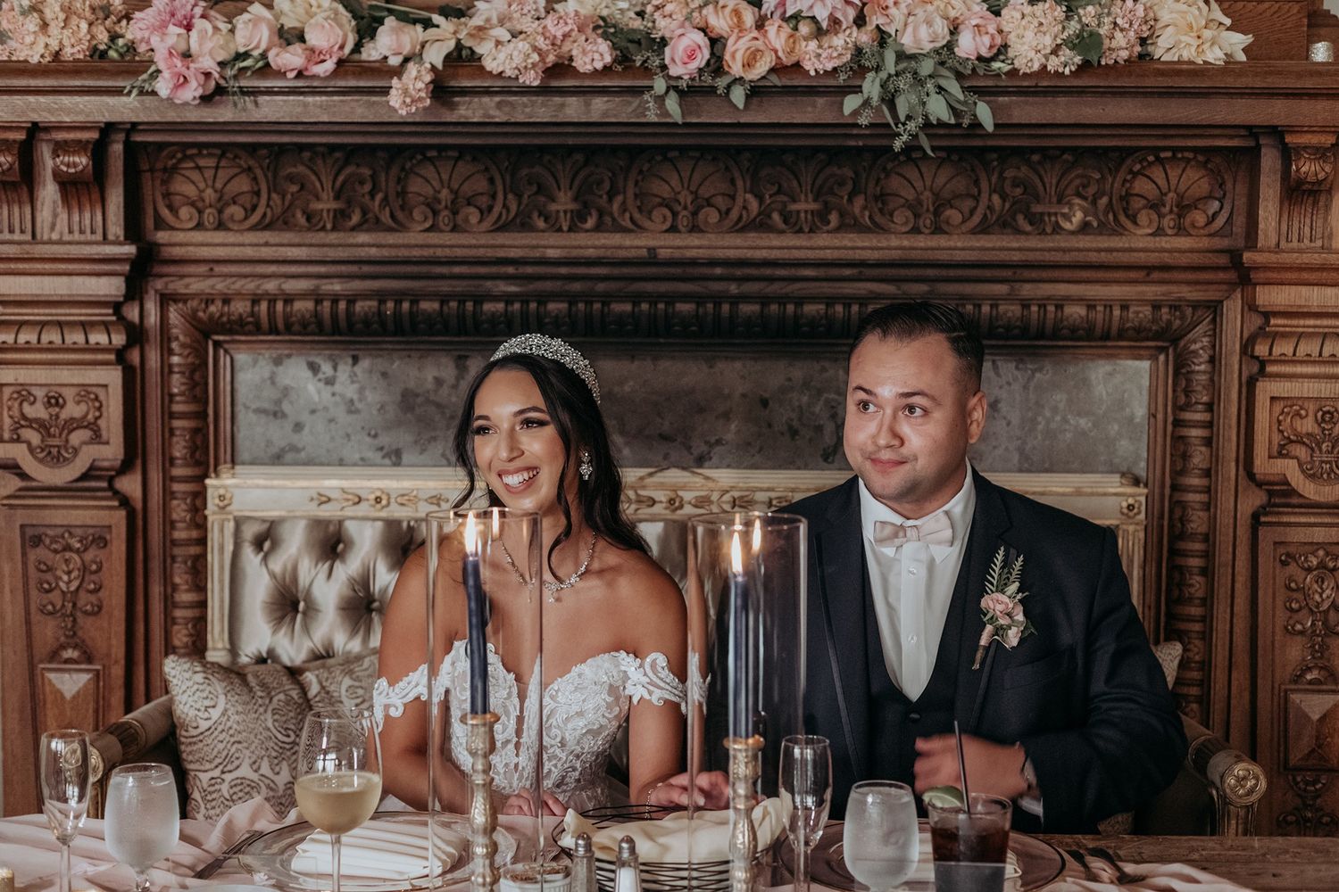 Bride and groom seated at elegant wedding reception table with candles and floral arrangements before ornate fireplace.