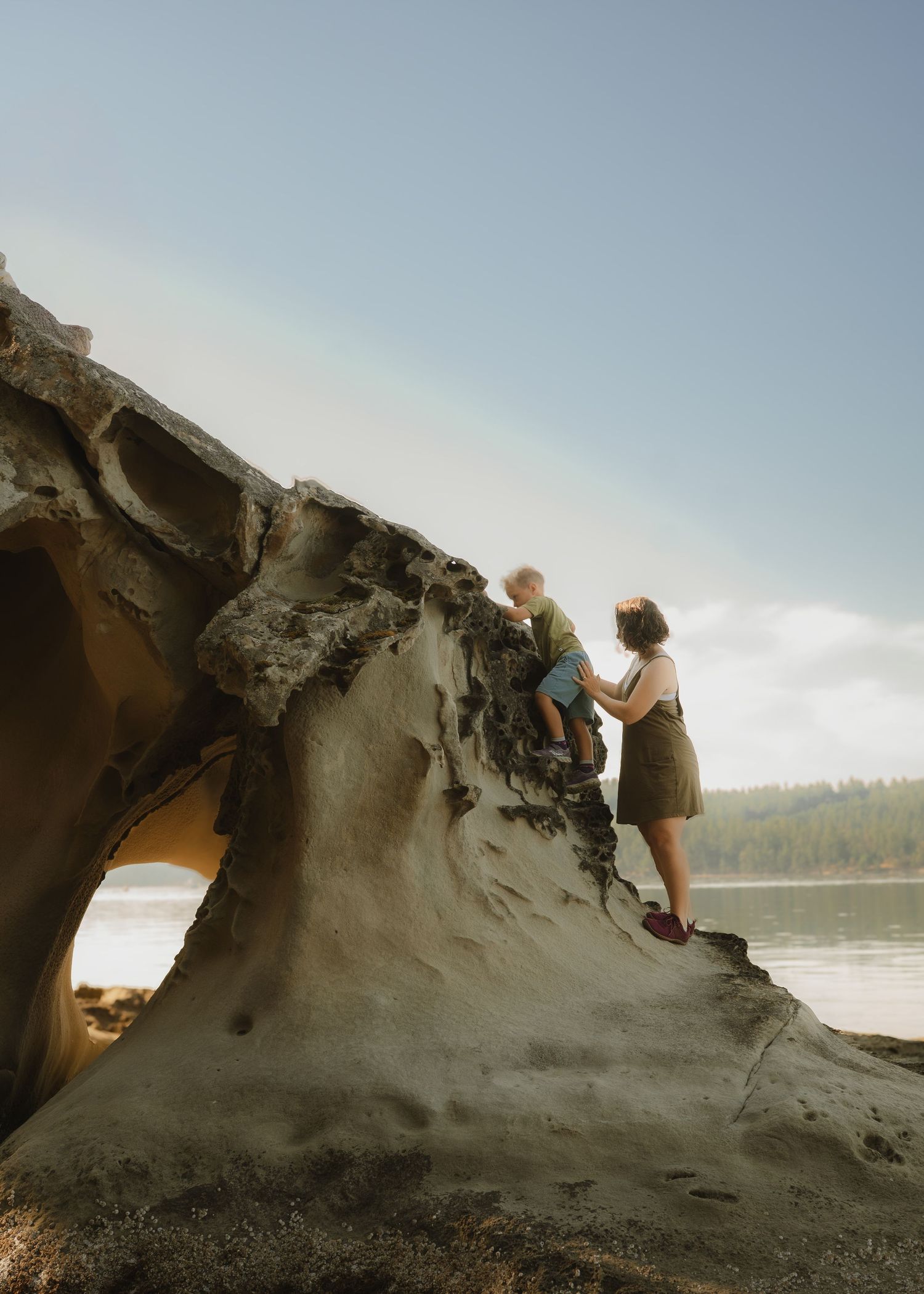 A woman helps a young child climb a dramatic eroded sandstone rock formation on a scenic beach with forest in the background.