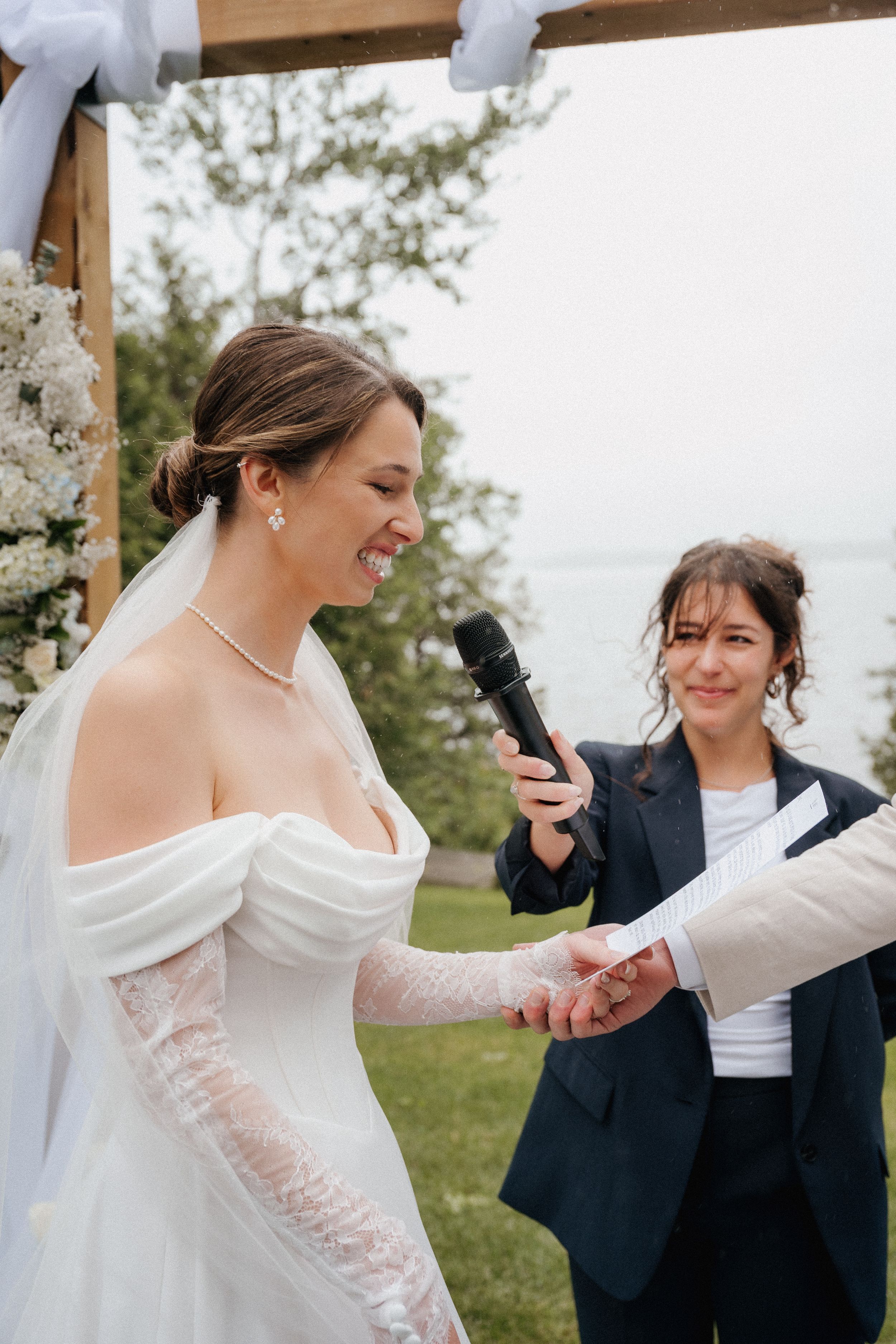 An outdoor wedding ceremony with a bride in a white lace gown and officiant holding a microphone by the water.