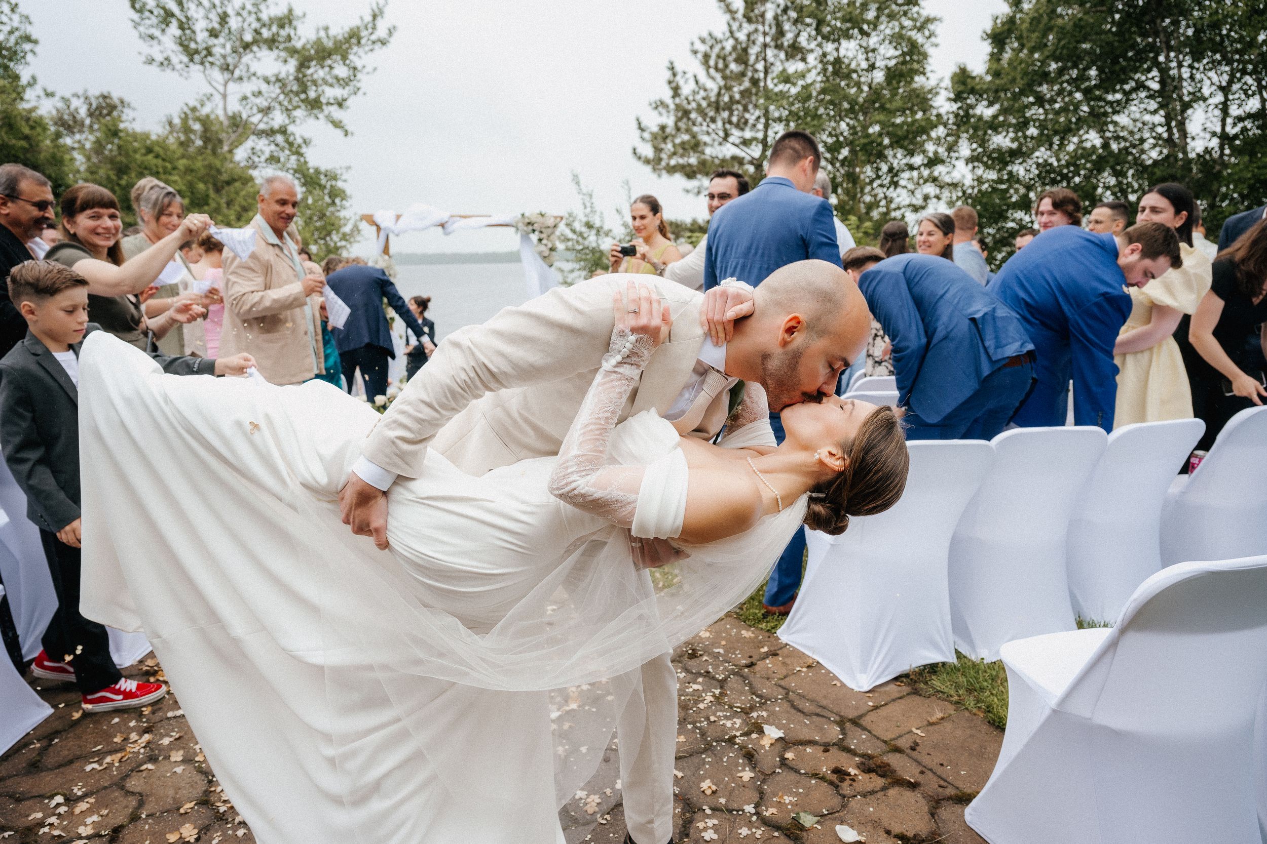 A romantic dip kiss during an outdoor wedding ceremony with guests and white chairs by the water.