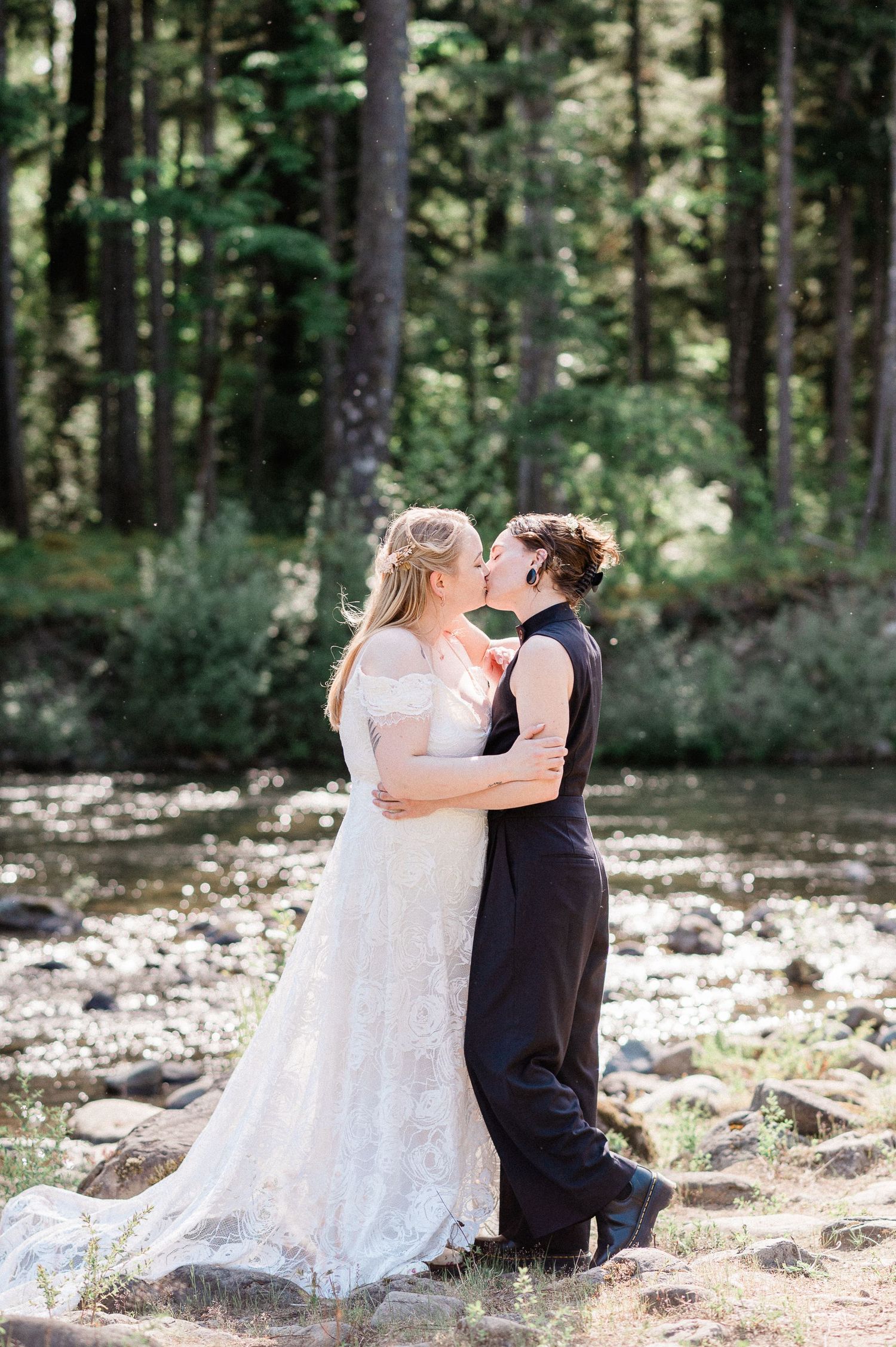 A romantic couple kiss on rocky ground in a sunlit forest setting with river behind them during their elopement.